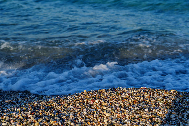 Close-up of small multicoloured pebbles on a beach with soft foamy wave washing ashore and blue sea in background.