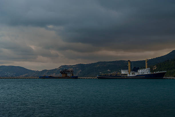 Two large cargo vessels lie moored along a breakwater with dark mountains and heavy storm clouds in the background.