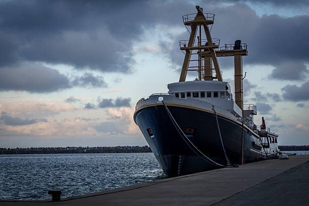 A large blue and white ship with tall yellow masts is tied along a concrete pier, its bow facing the camera as soft evening light glows beneath heavy clouds over the calm harbor.