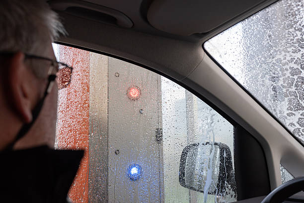 A man sits in the driver’s seat looking out the side window at the control lights of an automatic car wash, with water droplets visible on the glass.