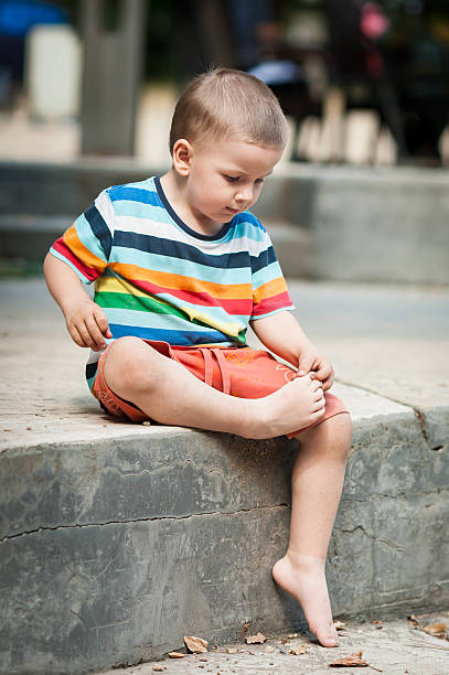 A young boy sits on a concrete ledge, wearing a colorful striped shirt and shorts. He is focused on playing with a stick, enjoying his time outdoors in a sunny setting.