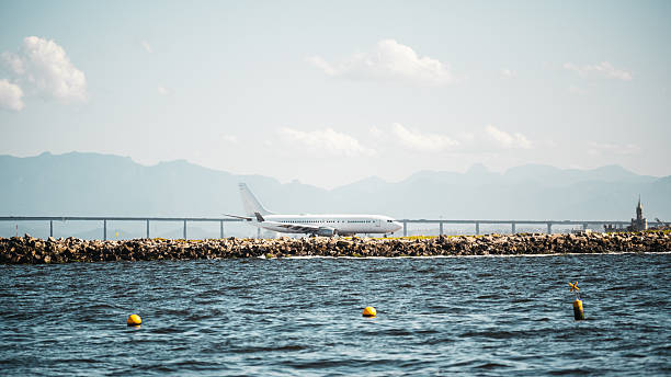 Commercial airplane taxiing along a rocky shoreline with distant mountains, calm sea water and bright marker buoys under clear sky in a long-lens telephoto coastal scene