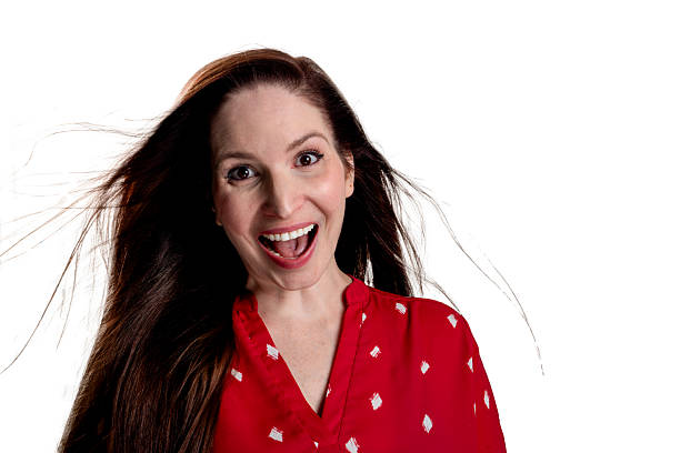 A young woman in a red polka dot shirt expresses extreme surprise with wide open mouth and wide eyes. Her long dark hair is dramatically windblown, suggesting sudden movement or emotional intensity against a white background.