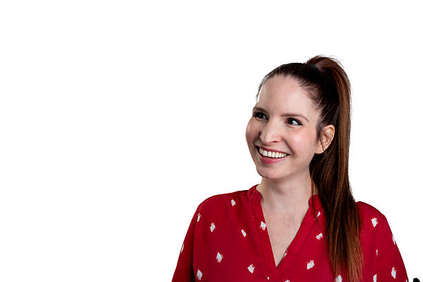 Young woman with long brown ponytail wearing a red blouse with white geometric print, looking off-camera with a wide, genuine smile. Clean white studio background provides clear contrast. Her hair is pulled back neatly, and her expression appears friendly and approachable.