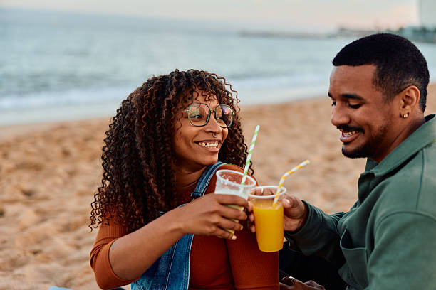 Young adult couple toasting refreshing drinks with striped straws, smiling and laughing during a romantic beach picnic on a sunny day, showcasing love and togetherness