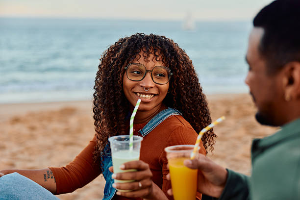 Young adult couple relaxing on a sandy beach at sunset, smiling and sharing refreshing drinks with straws while enjoying a romantic, carefree seaside moment together