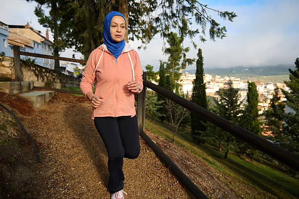 A Muslim woman in a peach jacket and blue hijab runs along a hillside path with a city view, portraying contemporary daily life, fitness, and independence in Europe.