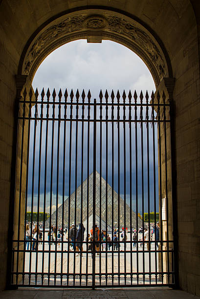 Paris, France - August 29, 2025: View from Paris shows the Louvre Pyramid framed by an ornate arch. Dark clouds gather above the busy courtyard. Tourists gather in the courtyard.