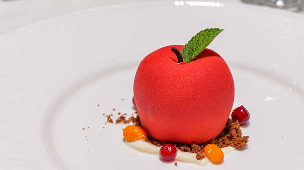 Red dessert shaped like an apple, garnished with mint leaf, surrounded by colorful fruit sauces and crumbled chocolate on a white plate