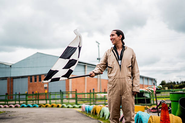 A jubilant race official stands at a go-kart track, holding a classic black and white checkered racing flag with enthusiasm. The industrial setting with colorful tires and buildings creates a vibrant backdrop for this motorsport moment.