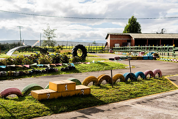 A vibrant go-kart racing track featuring a yellow podium with numbered positions, surrounded by creatively arranged colored tires used as track barriers. The circuit is set in a green, grassy area with a rural landscape, including a brick building and power lines in the background, suggesting a local recreational facility.