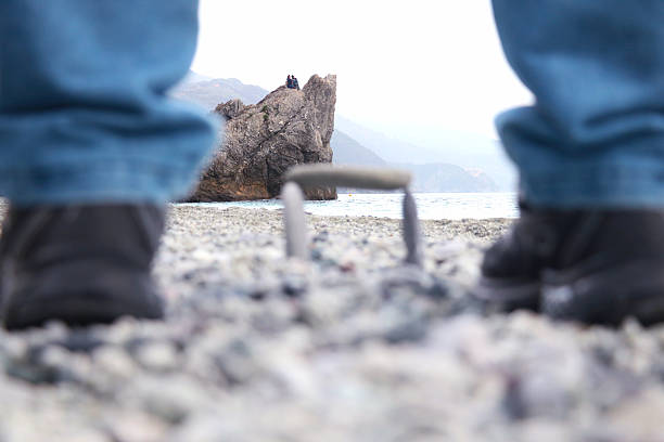 Creative low-angle shot on a rocky beach, framing a distant cliff where two people sit overlooking the sea. Foreground includes blurred legs and stones, adding depth and perspective to the coastal landscape.