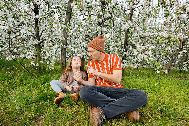 Father and daughter sitting together on the grass, enjoying a relaxing moment surrounded by blooming apple trees in an orchard.