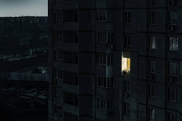 Close-up exterior shot of a multi-story Soviet-era brick apartment building at night, showing a single window brightly illuminated amidst the surrounding darkness caused by a power outage.