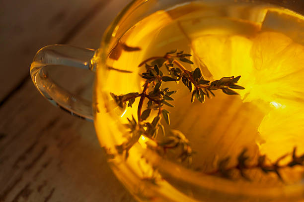 A glass cup of herbal tea with lemon and thyme on a wooden surface, highlighted by warm light and natural textures that evoke calm and wellness.