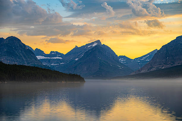 A stunning sunset illuminates the rugged peaks and calm waters of Glacier National Park in Montana. Soft mist floats above the lake as golden light paints the sky, creating a peaceful and awe-inspiring alpine landscape.