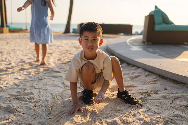 An Asian boy is looking at the camera and smiling while playing with sand on a tropical beach.