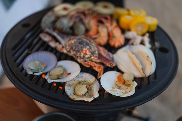 Assorted fresh seafood and corn are cooking on a portable grill outdoors.