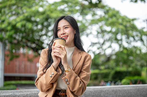 istock Pretty asian woman office worker holding coffee cup with both hands sitting on stone bench in garden 2247562598