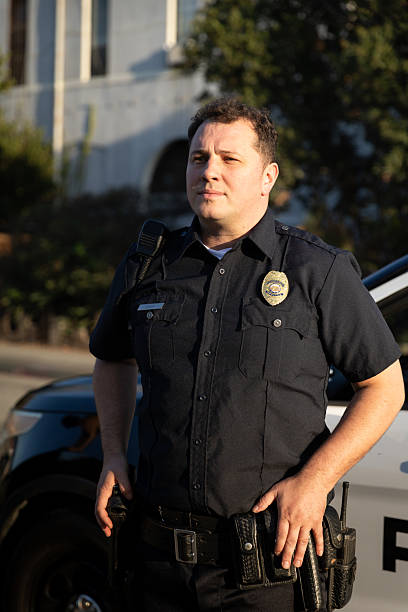 istock White Policeman With Hands on Belt Looks Off-camera While Posed in Front of Squad Car 2247565621
