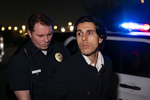 Shot of a police officer holding a criminal in handcuffs in front of a squad car at night.