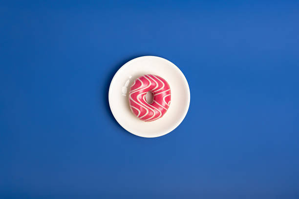 Tasty pink glazed strawberry donut on white round plate from above on blue background. Fast snack time.