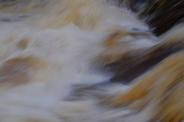 A blurred, abstract close-up of fast-moving water creates soft streaks of white, gold, and dark tones, capturing the energy and texture of a rushing stream in motion.