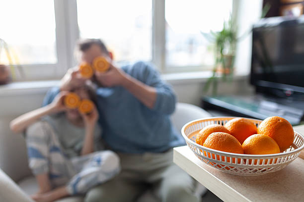 Basket full of oranges on kitchen countertop with blurred background. Man and boy holding two orange halves over eyes like goggles.