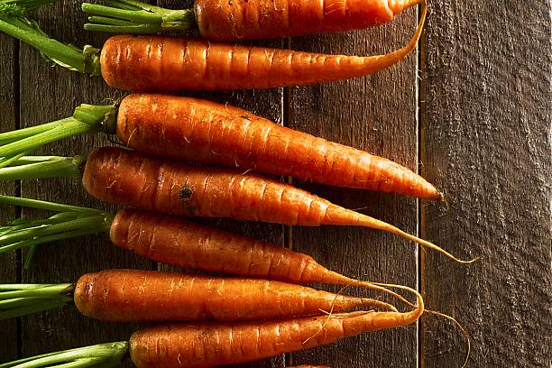 A vertical arrangement of vibrant orange carrots with green tops rests on weathered, dark wooden planks, showcasing their natural texture.