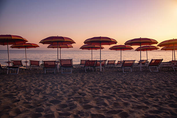 Calm sunset over a sandy beach with rows of red umbrellas and empty lounge chairs, inviting relaxation and vacation mood as calm waves meet the shore.