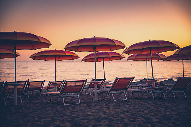 Calm sunset over a sandy beach with rows of red umbrellas and empty lounge chairs, inviting relaxation and vacation mood as calm waves meet the shore.