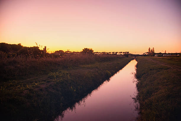 A tranquil sunset scene along a narrow canal, reflecting warm pink and orange skies. Grass and wildflowers line the banks, creating a peaceful, natural landscape perfect for nature or travel projects.