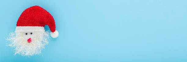 istock Santa Claus head with bright red hat and white beard on light blue table background. Pastel color. Closeup. Creating Christmas decoration. Wide banner. Empty place for text. Top down view. 2247601614