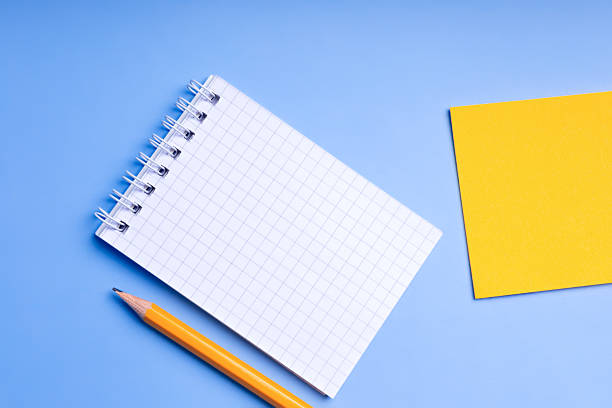Stationery from above. Checkered notepad mockup, orange pencil and paper for notes on blue background top view. Place for text.