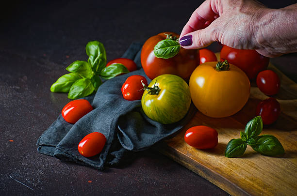 A hand places a basil leaf on a vibrant selection of heirloom tomatoes, including yellow, green, and red varieties. Arranged on a rustic cutting board with cherry tomatoes and fresh basil leaves. Natural light highlights the freshness and zero-waste approach to healthy, home-cooked meals. Copy space available.