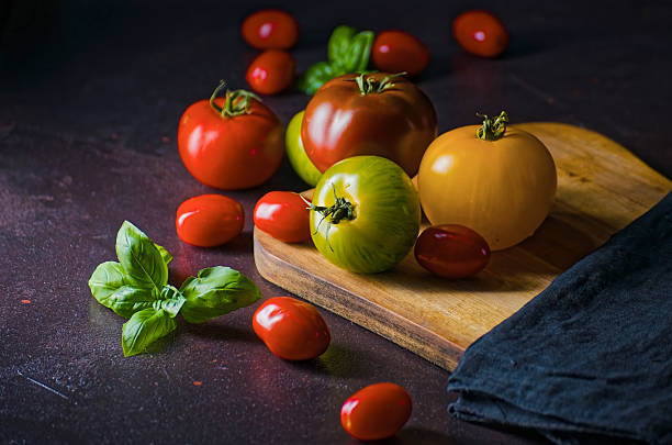 An assortment of ripe tomatoes including red, green, and yellow varieties rest on a wooden board with fresh basil. Designed with ample negative space for marketing materials or social media content promoting anti-inflammatory diets and healthy living.