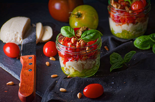 Layered salad in a glass jar featuring fresh tomatoes, pine nuts, creamy mozzarella, and green basil. A vibrant and healthy meal preparation on a dark textured surface with copy space. Ideal for concepts of healthy eating, Mediterranean diet, or meal prepping.
