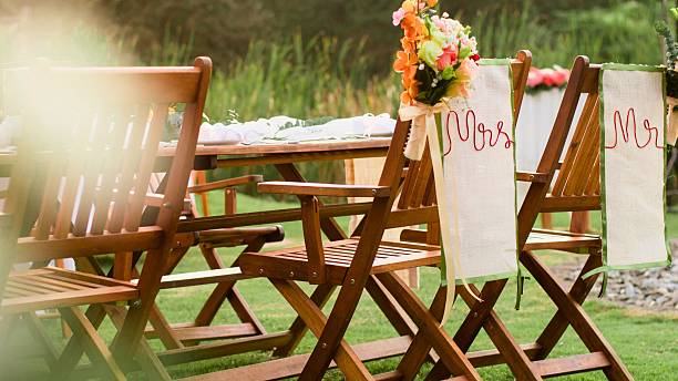 Wooden chairs are arranged for an outdoor wedding ceremony, adorned with floral arrangements and decorative banners.