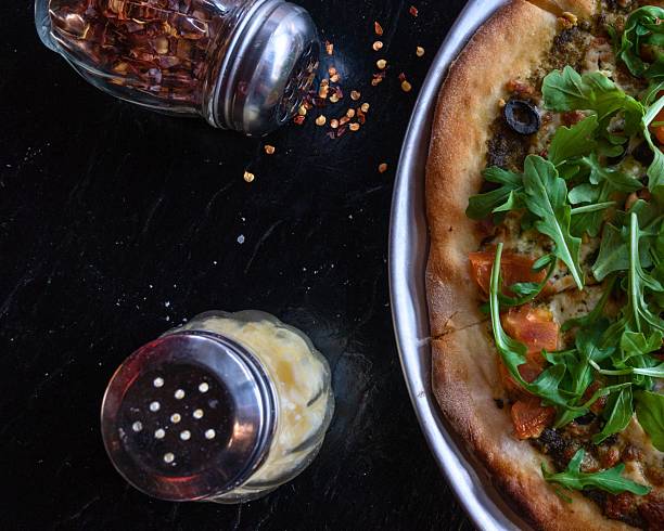 A close-up shot of a pizza with fresh green toppings and spices being sprinkled from shakers onto a dark surface.