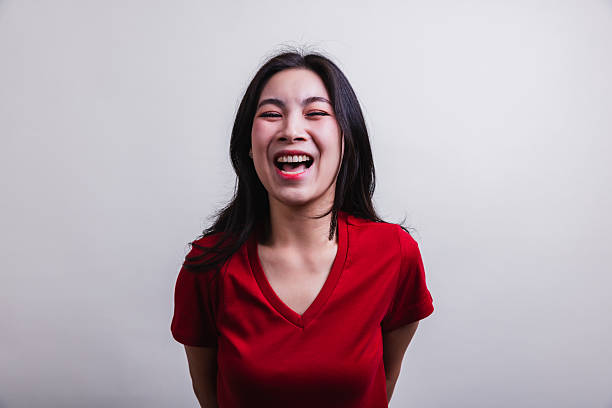 A young woman wearing a red shirt laughs heartily while standing against a simple light grey background. Her joyful expression radiates happiness and warmth.