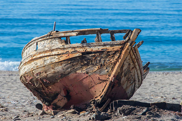 An old, weather-beaten boat rests on a sandy shore, its blue stripe peeling away. Rusty nails, rough wood, and scattered stones evoke nostalgia, solitude, and coastal history