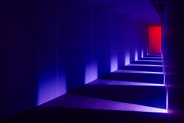 A futuristic corridor illuminated with blue and purple neon lighting, creating long geometric shadows along the floor and walls. The pathway leads toward a glowing red light, adding dramatic contrast and a mysterious sci-fi atmosphere. Perfect for themes of technology, virtual spaces, cyber environments, tension, and abstract architecture.