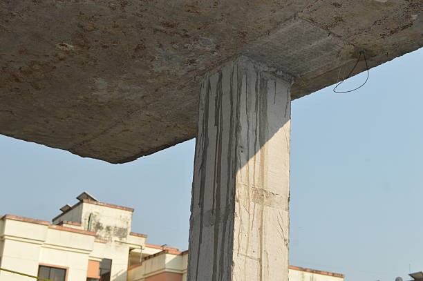 A concrete pillar supports a heavy slab at an urban construction site. Weathered stains streak the column while nearby modern buildings rise, highlighting industrial textures and city development.