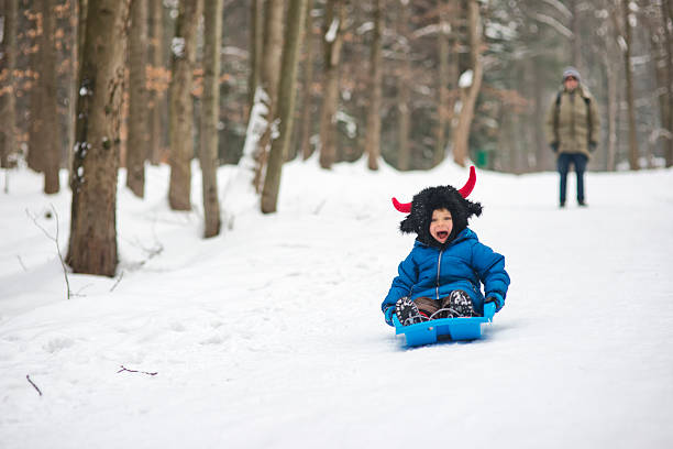 A child dressed in a blue winter jacket and a black furry hat with red horns sleds down a snowy path in a forest, mouth open in excitement. Seated on a blue sled, the child speeds past tall trees dusted with snow. Father stands in the background, watching the descent. The image captures a thrilling moment of outdoor winter play, blending costume humor with the exhilaration of sledding in a natural setting.Shot with Nikon D800.