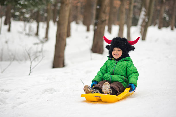 A child dressed in a green winter jacket and a black furry hat with red horns sleds down a snowy path in a forest, mouth open in excitement. Seated on a yellow sled, the child speeds past tall trees dusted with snow. Shot with Nikon D800.