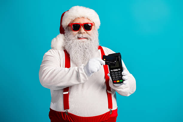 istock Santa with red suspenders and white beard pays with a card in a bright blue studio for festive season shopping and Christmas sales imagery 2247644572