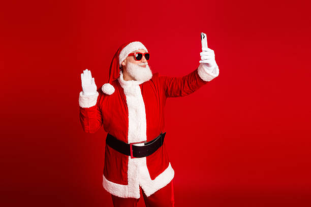 istock Santa selfie in red suit wearing sunglass while taking a festive self portrait against a bold red background 2247647475