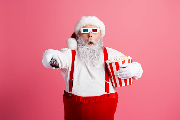 istock Santa with red suit and white beard holds popcorn bucket and points remote in a playful festive scene against a pink background 2247647792