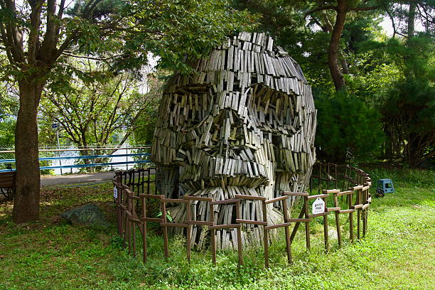 A large weathered wooden head sculpture rises behind a low fence in the shaded grove at the Animation Museum park in Chuncheon.