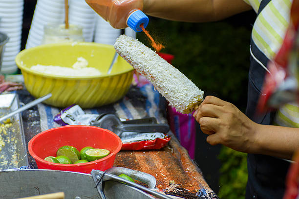 A street vendor prepares corn on the cob with various condiments. Elote preparation.